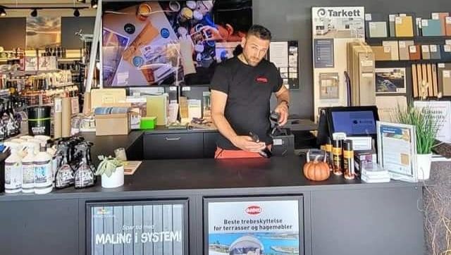 Ansatt bak disk i malingsbutikk, omgitt av produkter og reklamemateriell fra ulike merker.