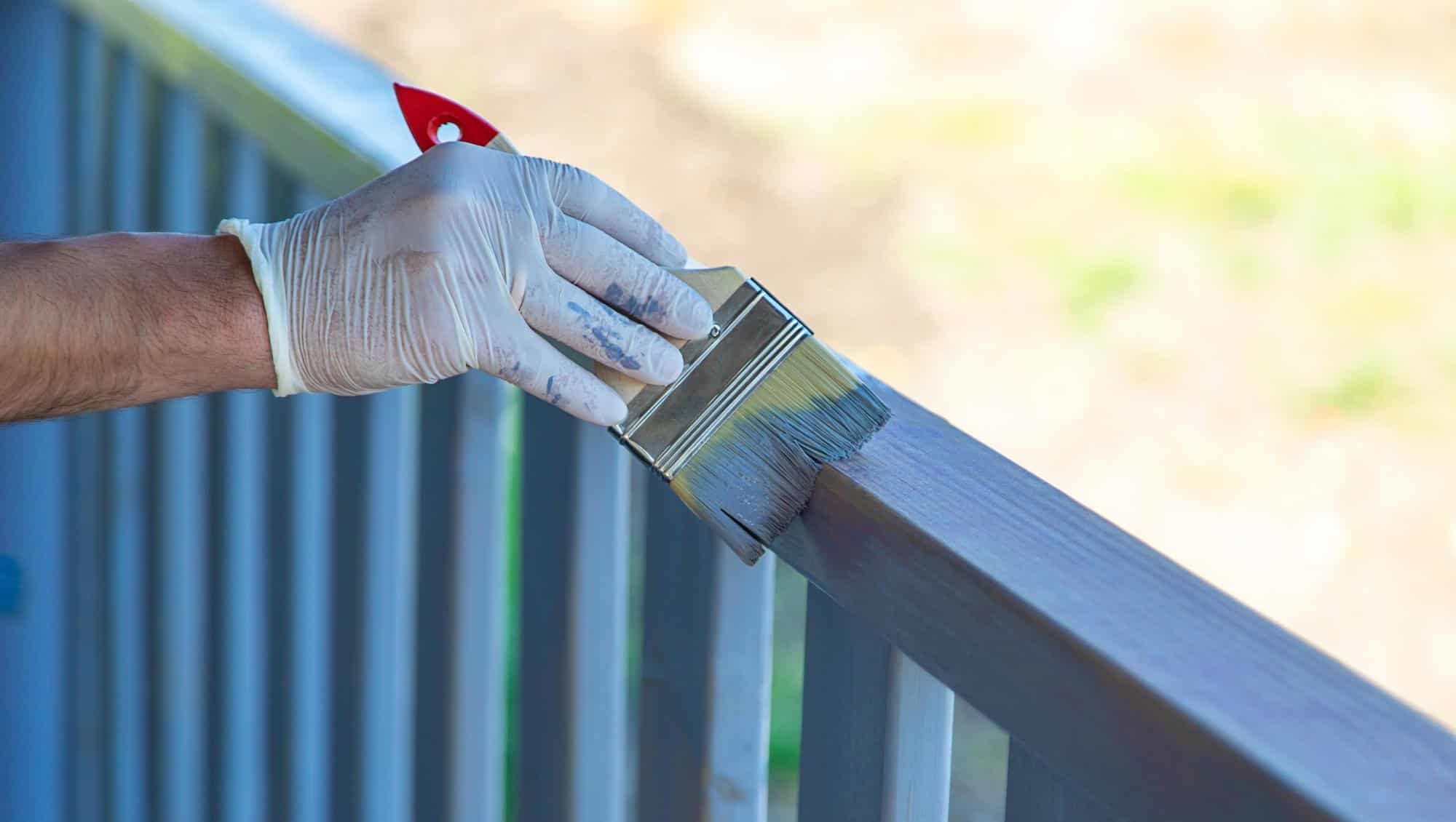 Hånd med hanske maler et utendørs rekkverk blått med pensel, i naturlig dagslys.