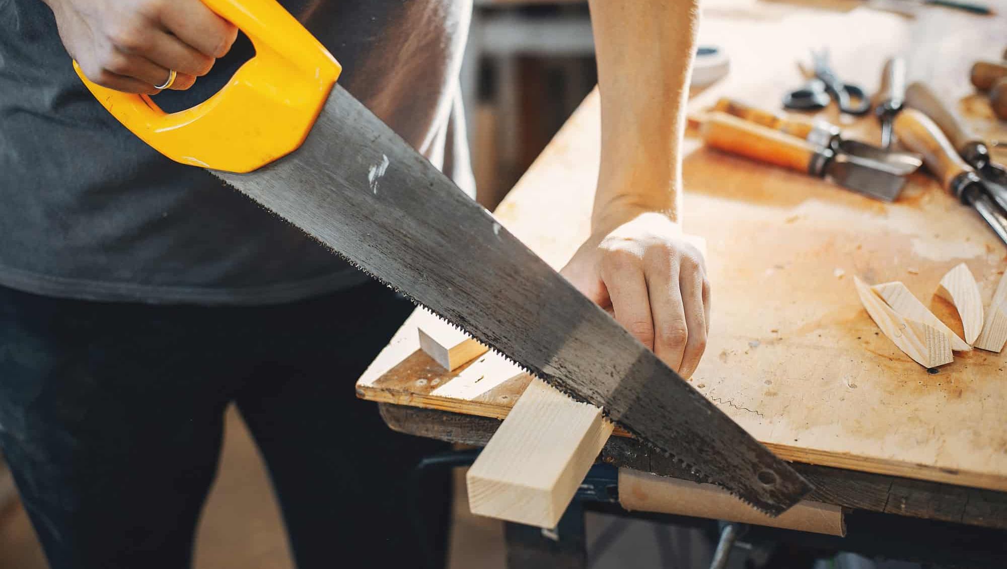 Person sager et stykke treverk med håndsag på arbeidsbenk omgitt av ulike treskjæringsverktøy