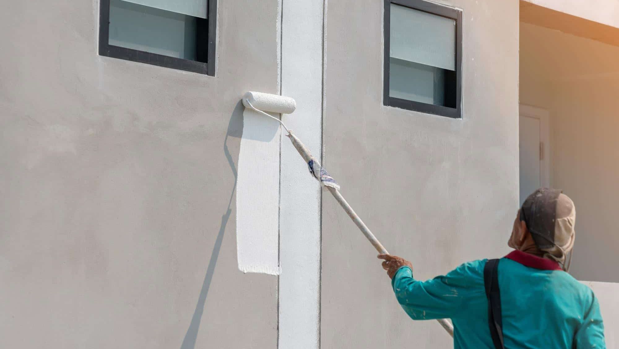 En person maler en fasade med hvit maling ved hjelp av en rulle festet på et langt teleskopskaft.